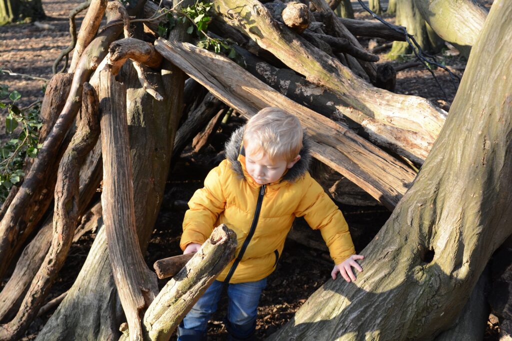 white toddler in yellow winter coat and jeans holding a stick in a wooden den