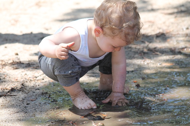 white toddler in grey trousers and white vest crouched an squishing mud