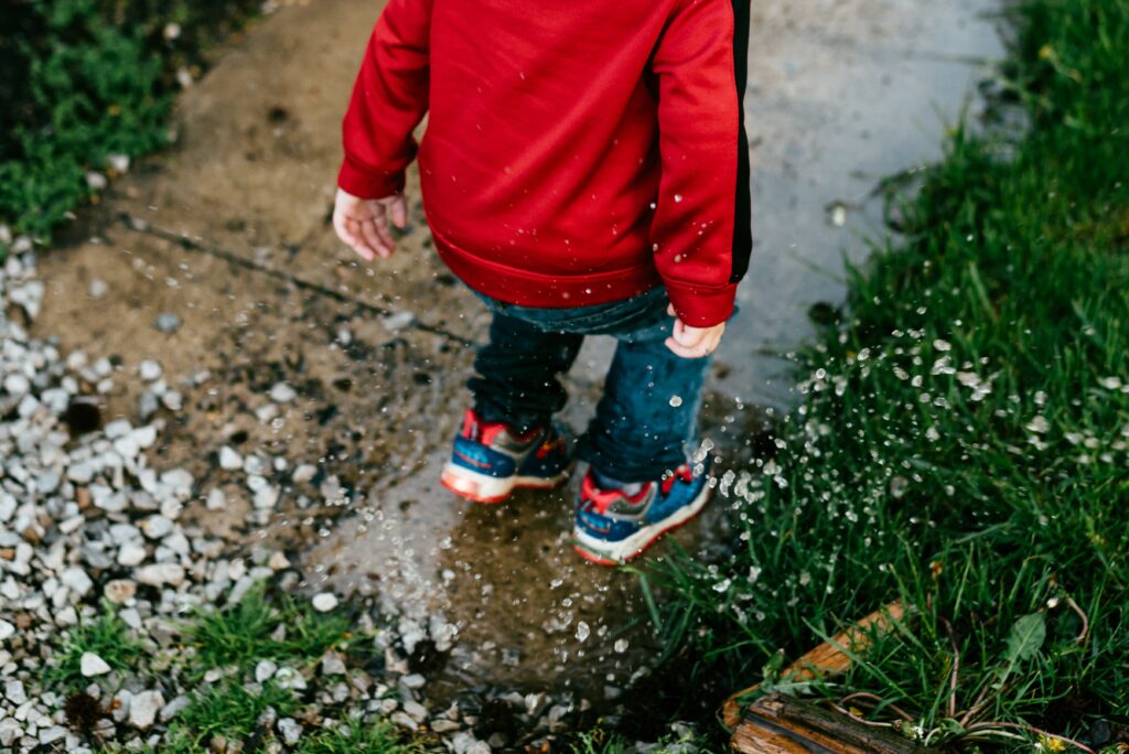 toddle in jeans, trainers and red hoodie splashing in a puddle